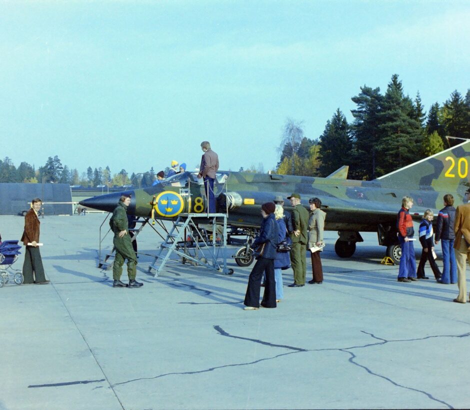 Ett kamouflage-målat flygplan, stående på en flygplats eller flygfält. Civilklädda människor står runt omkring det och tittar.