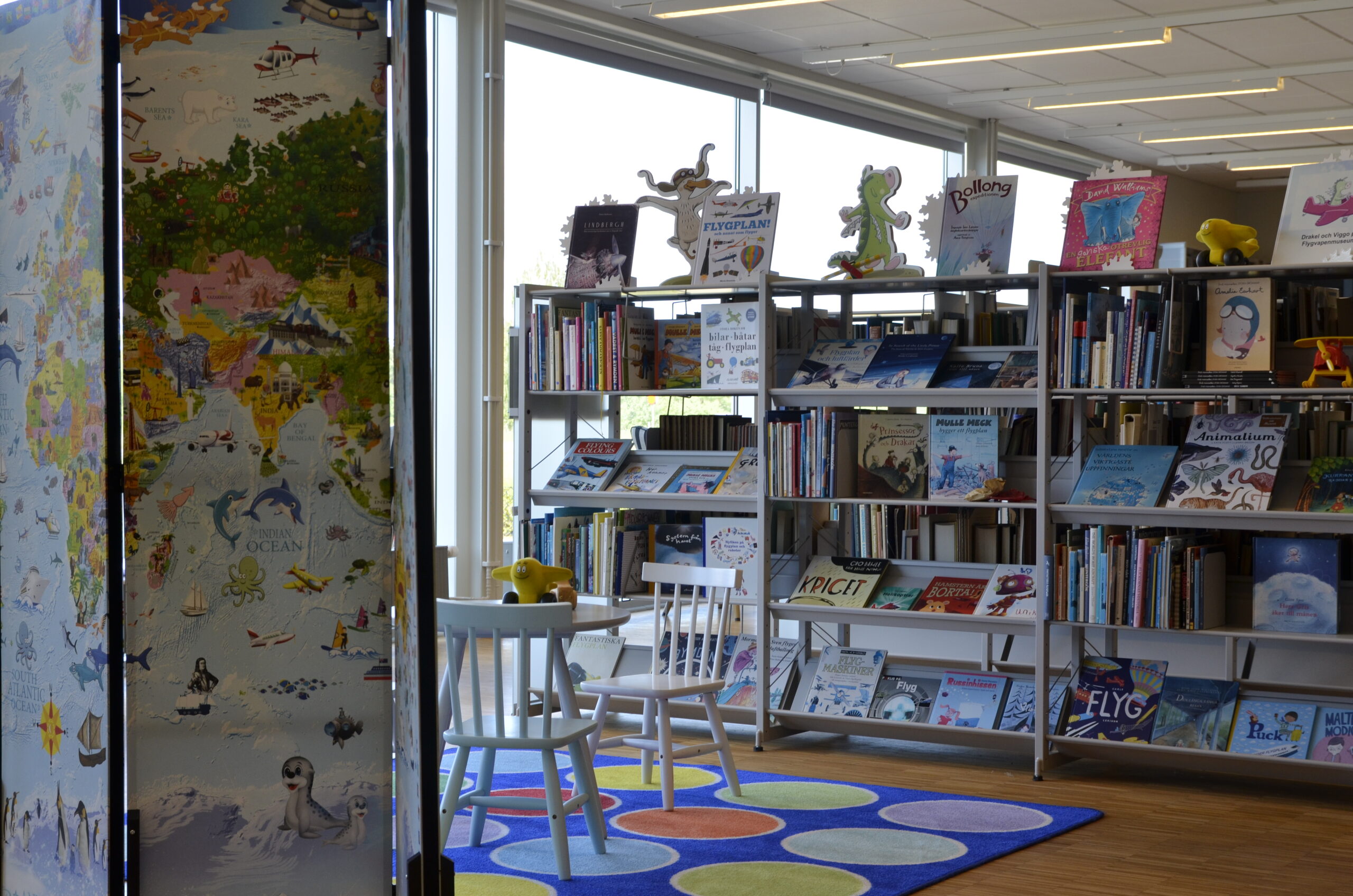 I bibliotekets barnhörna står bord och stolar som omges av hyllor med barnböcker.